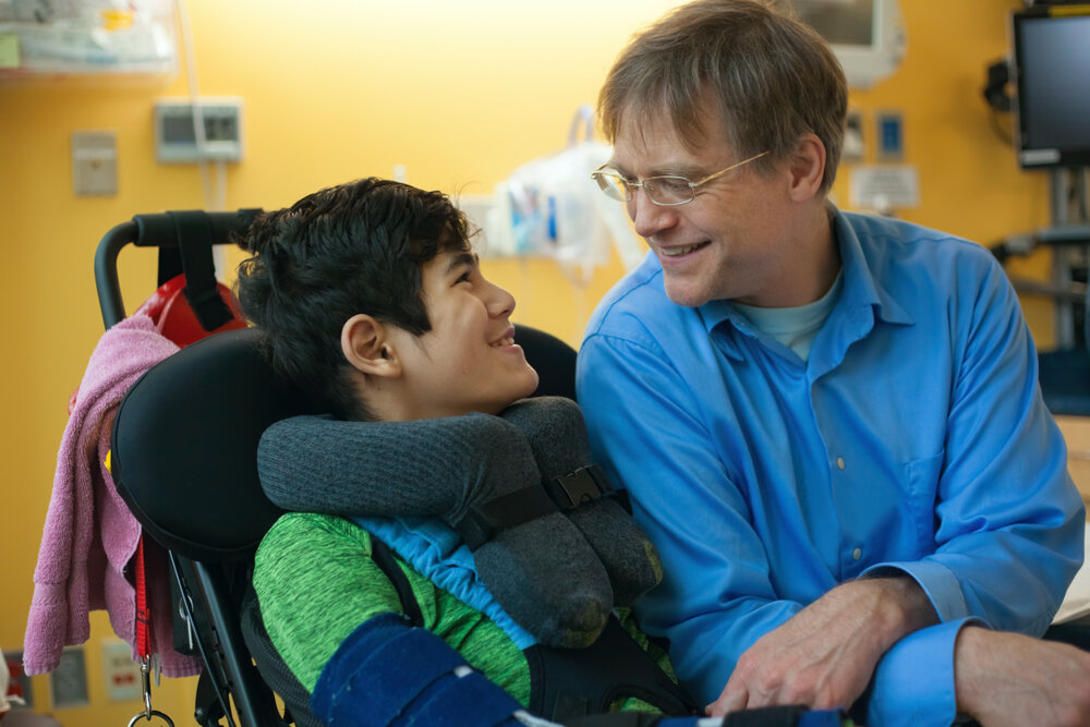 Hospitals in Europe offer World Class Pediatric Care. Smiling Father Sitting Next To Disabled Son In Wheelchair.